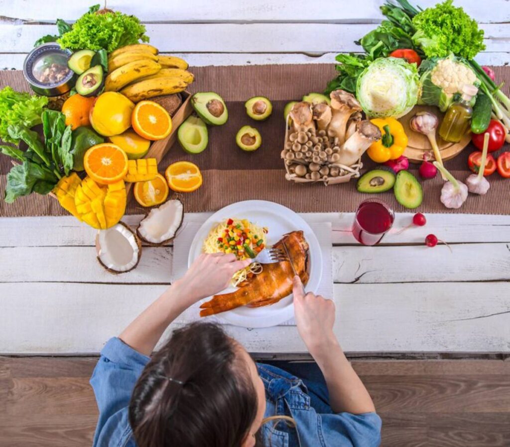 Top view of a woman eating a plate of fish surrounded by an assortment of fresh fruits and vegetables on a wooden table.