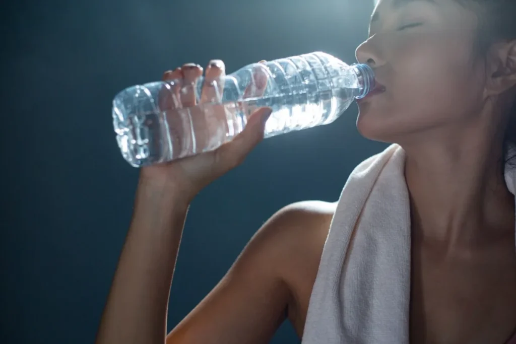 A woman drinking water from a bottle, with a towel hanging over her shoulder.