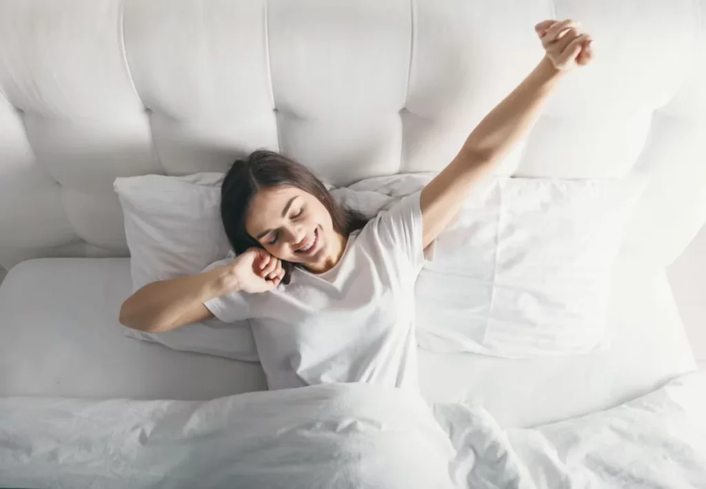 Young woman stretching happily in bed after waking up, surrounded by white bedding and pillows.