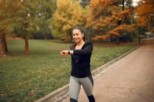 A woman in workout gear checking her watch in a park with autumn trees.