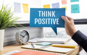 A person holding a "Think Positive" sign in a bright, organised office space with a clock, notebook, and sticky notes.