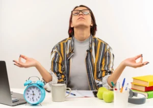Woman meditating at desk with laptop, books, and an alarm clock, seeking calm amidst work stress.