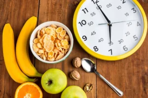 Healthy breakfast items including corn flakes, bananas, apples, orange, and nuts displayed with a large wall clock.
