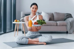 Young woman in sportswear holding vegetables and juice, seated on yoga mat in a modern living room.