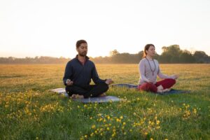 Two people meditating on yoga mats in a field at sunrise, surrounded by yellow flowers.