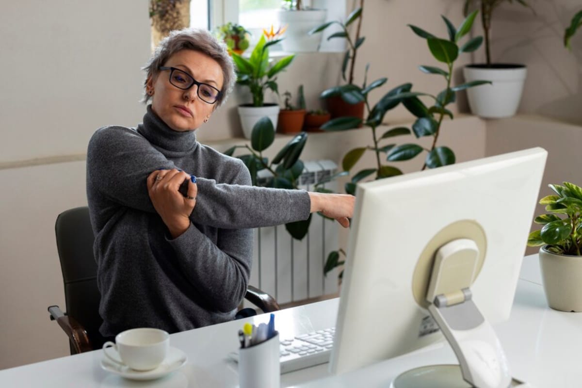 A woman with glasses stretches her arm while sitting at a desk with a computer and houseplants.