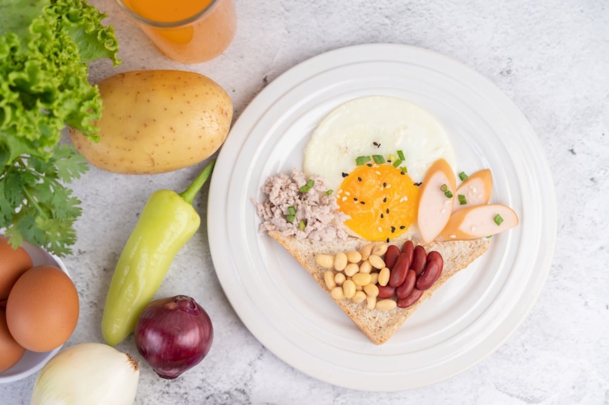 Plate with a creative breakfast arrangement, featuring toast, eggs, sausage, beans, and garnishes, with fresh vegetables and orange juice on the side.