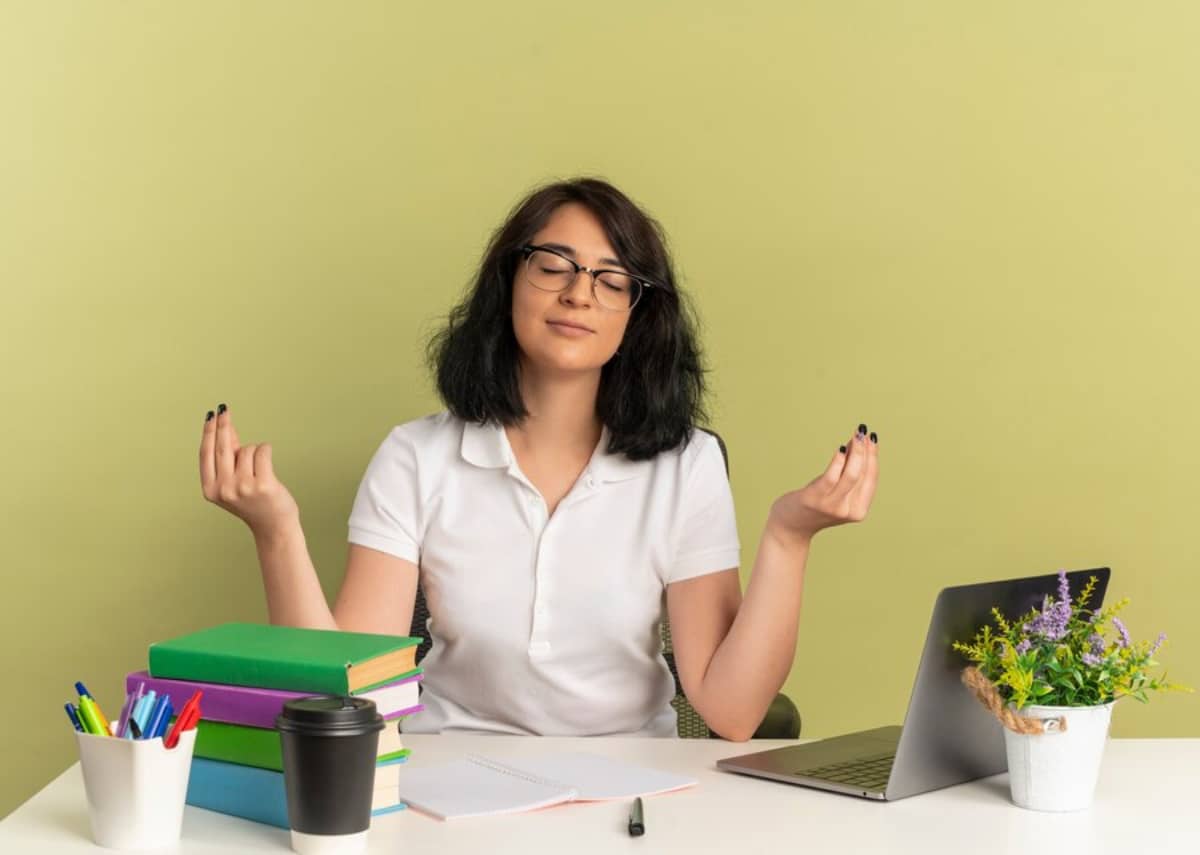  Young woman meditating at desk with laptop, books, and coffee, in a serene office setup.