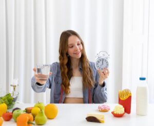 Young woman holding a glass of water and a clock, comparing healthy and unhealthy food options on a table.