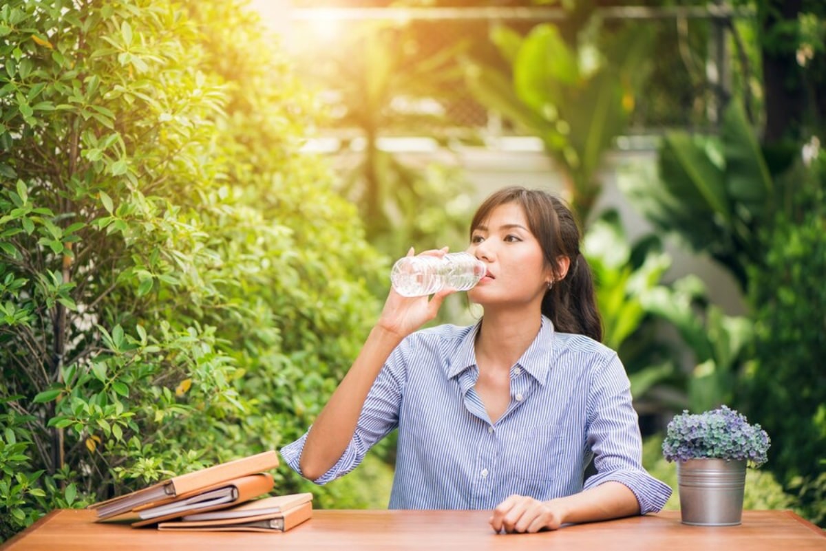  Woman drinking water from a bottle while sitting at a table outdoors surrounded by lush greenery.