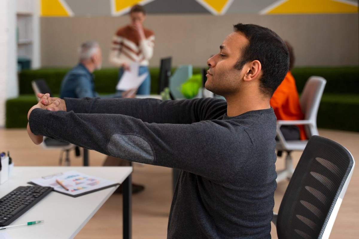 Man stretching his arms in an office environment, his colleagues blurred in the background.